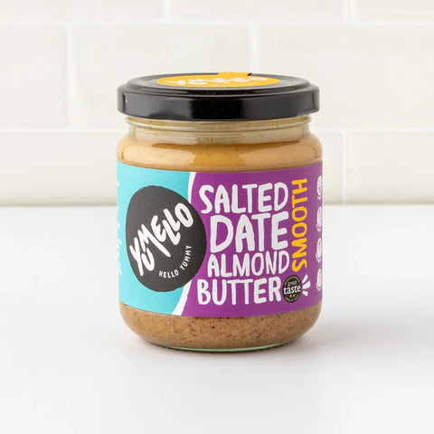 A jar of Yumello Salted Date Almond Butter on a white surface with a white tiled background. The jar has a black lid and a colorful label with purple, teal, and black sections