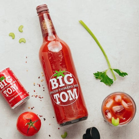 Bottle of Big Tom Spiced Tomato Mix with a can, tomato, and cocktail on a light background