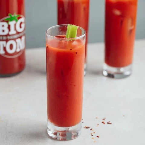 Glass of Bloody Mary with celery stick next to a bottle labeled 'Big Tom' on a light surface.