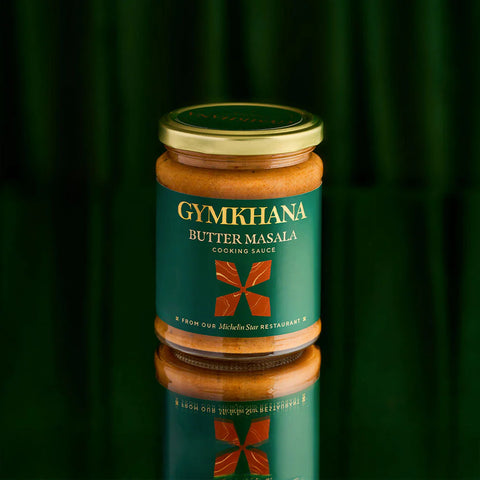 A glass jar of Gymkhana Butter Masala Cooking Sauce with a gold lid and a dark green label, set against a dark green background with a clear reflection below.