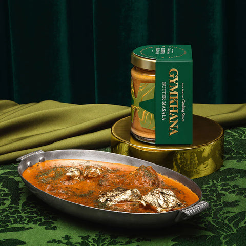A jar of GYMKHANA Butter Masala sauce in a green box sits on a gold pedestal next to an oval silver plate filled with the prepared orange-red curry, set on a green patterned cloth with olive drapes in the background.