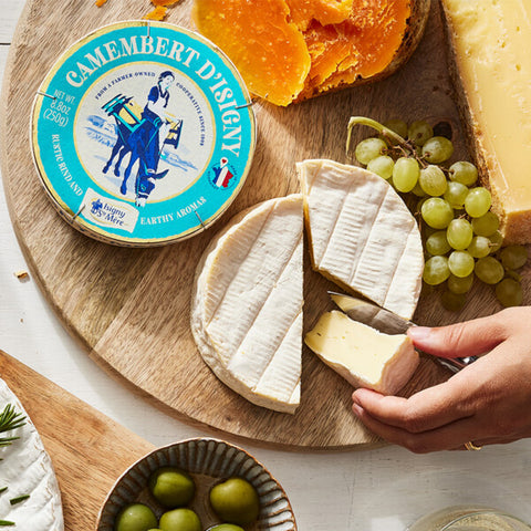 A top-down view of a cheese charcuterie board featuring a round container of Camembert d'Isigny, sliced soft cheese, green grapes, orange slices, and an olive, with a hand holding a knife to cut a piece of cheese.