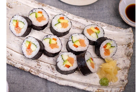 A serving of sushi maki rolls filled with cucumber, avocado, and salmon, arranged on a rustic white ceramic rectangular plate with ginger and wasabi garnish.