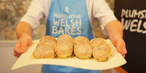 A person wearing a blue apron that reads 'PLUMSTONE WELSH BAKES' holds a white plate with six golden-brown baked pastries dusted with powdered sugar in a kitchen setting.