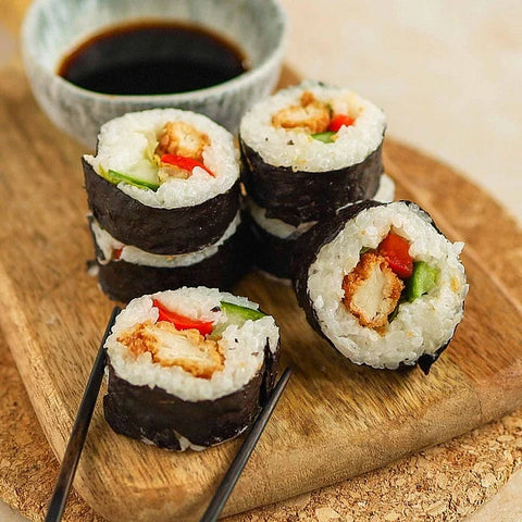 Four pieces of maki sushi rolls arranged on a wooden cutting board, with a small bowl of dark sauce in the background and black chopsticks at the side.