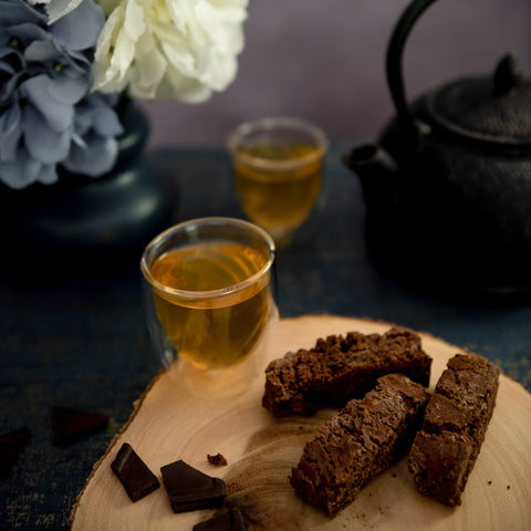 Three chocolate biscotti cookies on a round wooden serving board next to two small glasses of tea and dark chocolate pieces, with a black teapot and flowers in the softly blurred background.