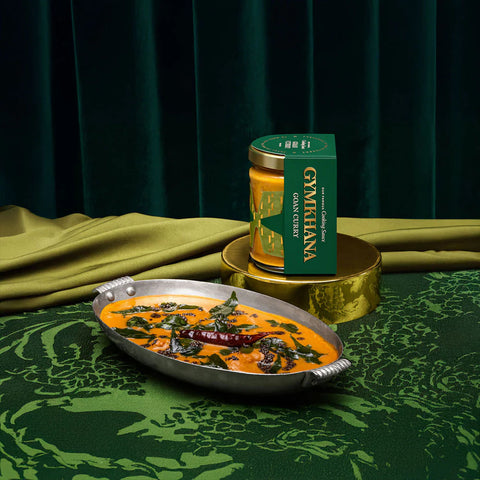 A jar of Gymria Organic Curry Paste sits on a gold stand next to a plated cooked curry dish, set against a dark green curtain and patterned green fabric backdrop.