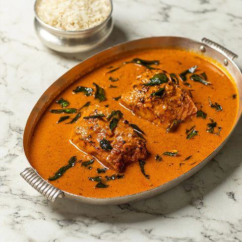 A serving of chicken in a rich orange-red curry sauce with chopped green leaves, presented in a silver oval metal dish on a white marble surface, with a small bowl of white rice in the background.