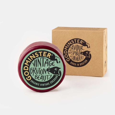 A round tin of Godminster Vintage Bruton Beauty organic vintage cheddar next to its brown kraft paper packaging box, both featuring black and light green branding on a clean white background.