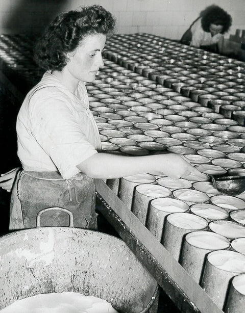 Black and white photograph of a woman working in a camembert factory, placing items into rows of metal canisters, with another worker in the background.