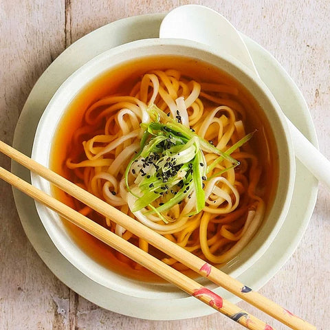 A top-down view of a bowl of noodles with yellow noodles in an orange-brown broth, topped with green scallions and black sesame seeds, served with wooden chopsticks and a white spoon on a light-colored wooden table.