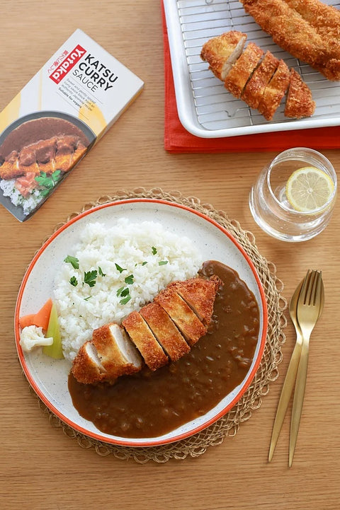food on a plate next to cutlery and a glass of water with a lemon and a box of Katsu Curry sauce