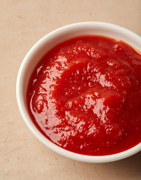 A close-up shot of a white ceramic bowl filled with bright red tomato sauce, set against a neutral beige background.