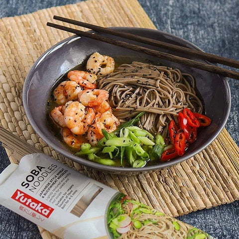 A bowl of soba noodles with shrimp, green onions, red chili peppers, and sesame seeds, served on a woven placemat with chopsticks and a tube of Yutaka Soba Noodles visible in the foreground.