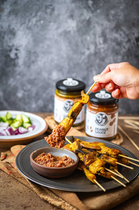 Person dipping a skewer into a bowl of satay sauce with jars of peanut butter in the background.
