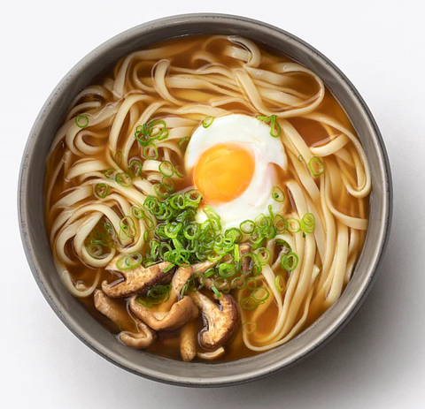 Top-down view of a bowl of udon noodles in a gray bowl, topped with a sunny egg, sliced green onions, and mushrooms, on a white background.