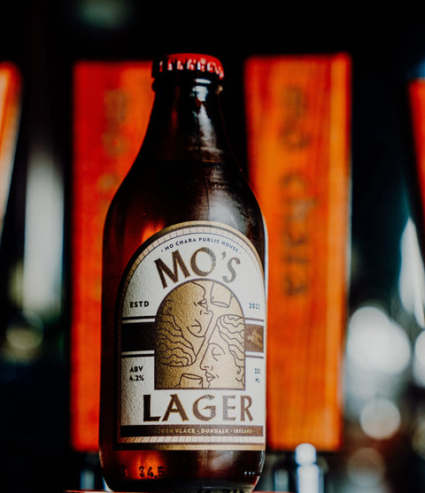 A bottle of Mo's Lager beer centered in the foreground with a blurred bar-like background featuring orange signs.