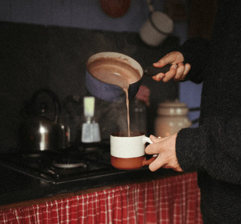 hand pouring hot chocolate out of a blue pot into a white and red mug in a kitchen