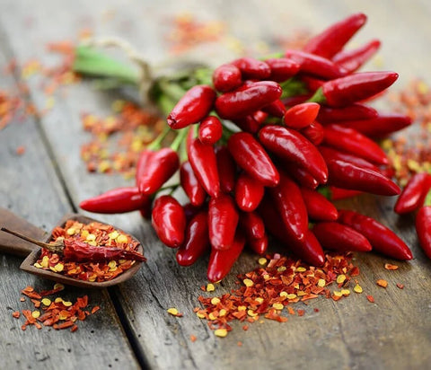 A cluster of bright red chili peppers with green stems, arranged on a rustic wooden surface with scattered red chili flakes and a small wooden spoon filled with chili powder.