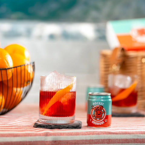 A cocktail glass filled with a red-orange drink and an orange slice sits on a table beside a small can labeled Pocket Hero, with oranges in a wire basket and a picnic-style basket blurred in the background.