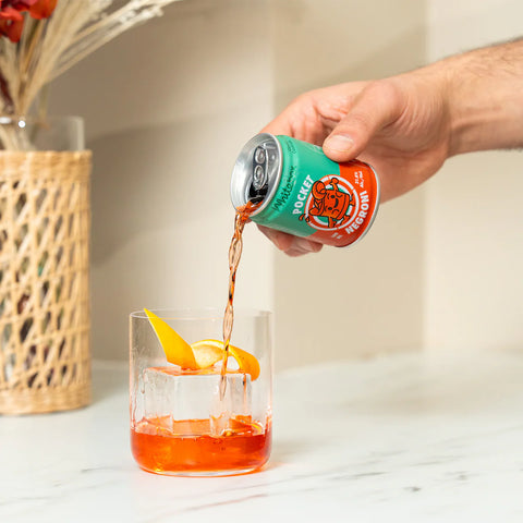 A hand pouring a green and orange can of White Box pocket negroni into a clear glass with ice, garnished with orange slices, on a white marble countertop.