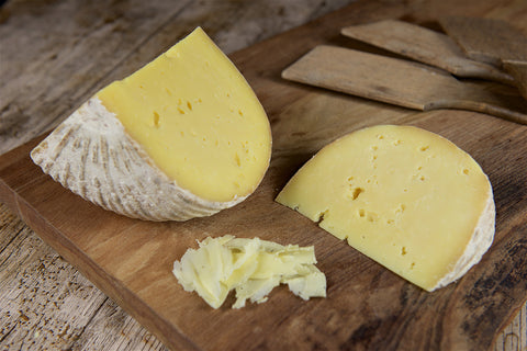 Two wedges of pale yellow cheese with textured off-white rinds on a wooden cutting board, with a small pile of shredded cheese in front and wooden cheese knives in the background.