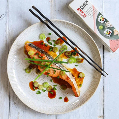 A plated salmon fillet with orange sauce, green onions, and sliced scallions on a textured white plate, with black chopsticks resting across it and a box of Wasabi Paste visible in the upper right on a light wood surface.