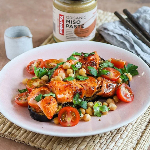 A plated salmon and tomato miso paste dish on a woven mat, with a jar of Yumeka Organic Miso Paste in the background.