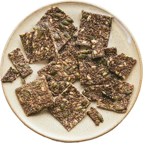 A top-down view of square gluten-free crackers topped with seeds and pumpkin seeds, served on a round beige ceramic plate against a white background.