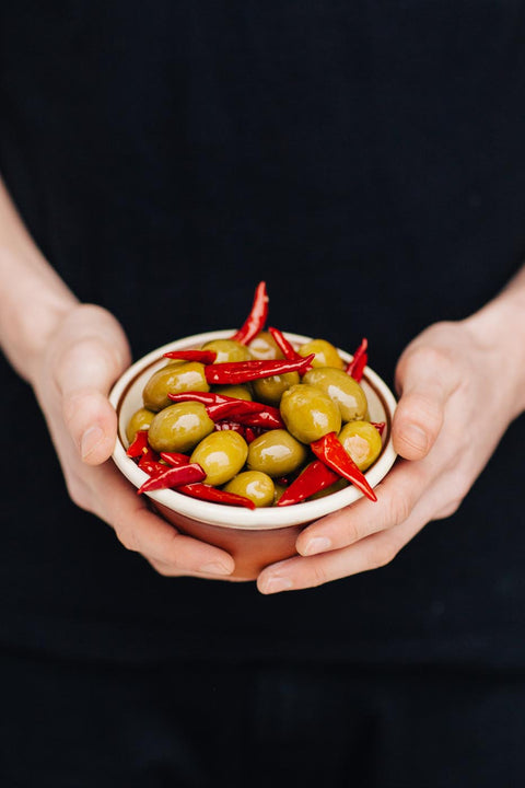 A close-up of a person's hands holding a small white bowl filled with green olives and sliced red chili peppers, set against a dark black background.