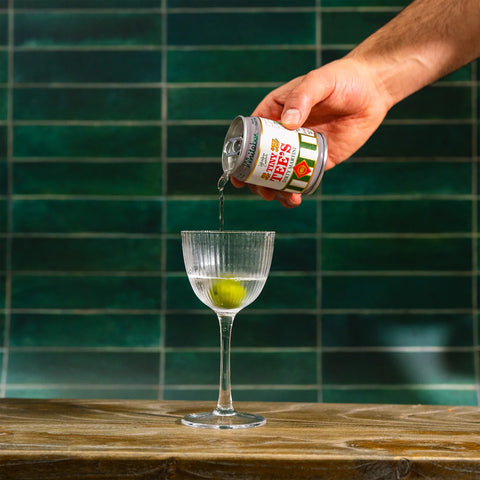 A hand pours a clear liquid from a can of tiny tee's dirty martini into a coupe glass containing a green lime wedge, set against a dark green tiled wall and a wooden counter.