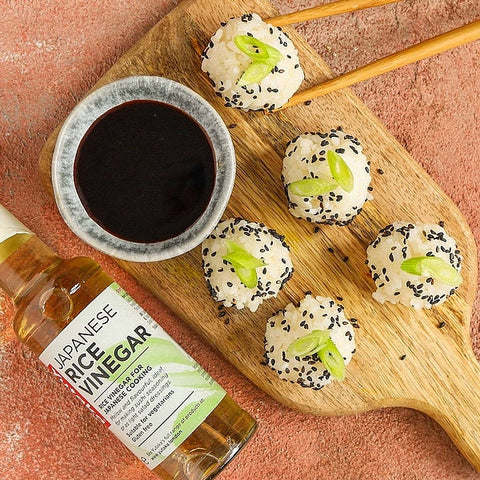 A bottle of Japanese rice vinegar with a green and white label is styled on a terracotta surface next to a small bowl of dark soy sauce and four sushi balls topped with black sesame seeds and green onions, arranged on a wooden board with chopsticks.