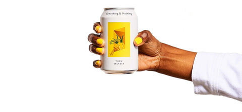 A hand with yellow nail polish holds a white can of Something & Nothing Puro Red Tea against a solid white background.
